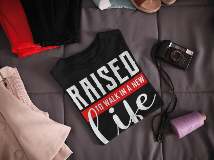 raised-to-walk-in-a-new-life-shirt-folded-t-shirt-mockup-lying-next-to-a-camera-and-clothes-on-a-bed